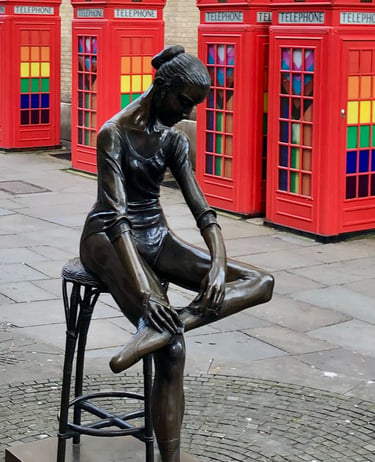 Bronze ballet dancer statue in London with red telephone boxes and colorful glass windows.