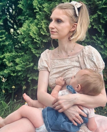 Mother sitting in front of a green bush, wearing a bow in her hair, holding and nursing her baby & looking away peacefully