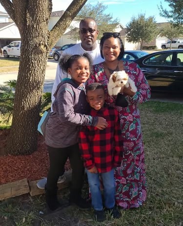 A happy family of four with their small white Mal-Shi puppy posing outdoors near a large tree.