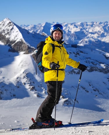 alt= "Xaver Kröll in einer gelben tobe Jacke und schwarzen Hose steht auf einem verschneiten Berg"