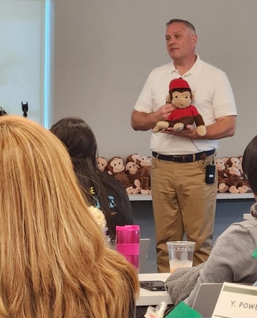 A man in a white polo shirt presents to a group while holding a Curious George plush toy.