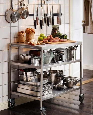 A mobile stainless steel table with three shelves used for food prep and storing kitchenware against a tiled wall.