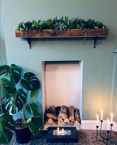 Rustic indoor fireplace featuring a succulent wooden mantle shelf, monstera plant, and lit candles.
