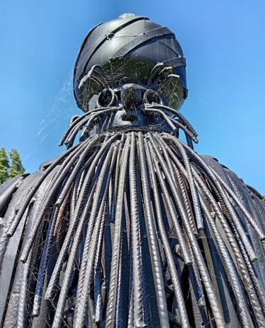 Black metal garden sculpture with a rebar beard and spider webs against a blue sky.