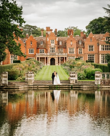 Bride and groom standing by a lake in front of a grand manor house