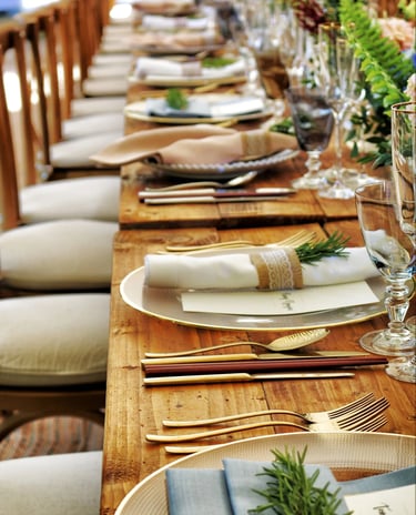 Rustic wedding table setting with gold cutlery, glass chargers, and rosemary sprigs on a wood farm table.
