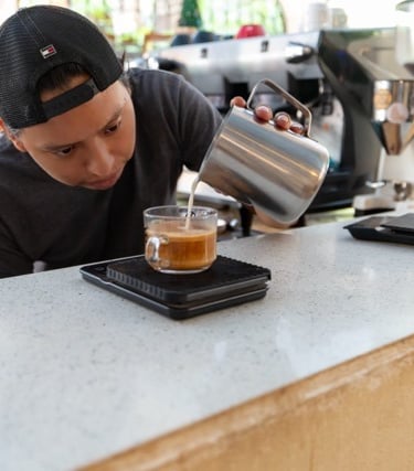 A barista pouring steamed milk into a glass of espresso to create a cappuccino on a coffee scale.