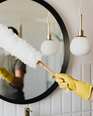 Cleaning a mirror with care — a gloved hand holding a duster in a bright, clean bathroom