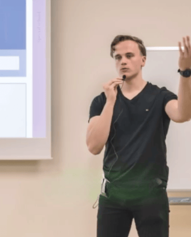 Julius-Sensei steht in einem Klassenraum vor einer Projektionsfläche, hält ein Mikrofon und erklärt gestikulierend ein Thema