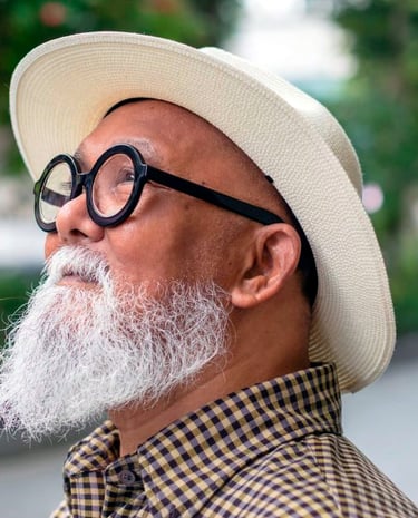 Stylish senior man with a white beard, round glasses, and a cream fedora hat looking up.