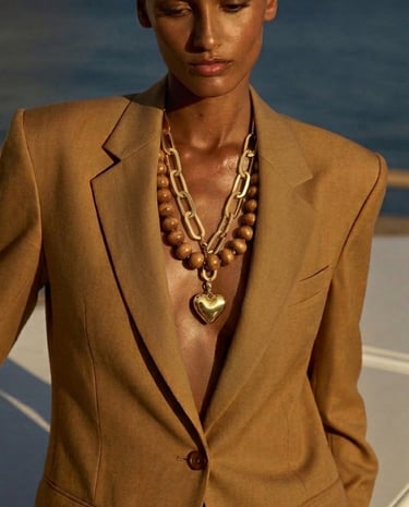 A woman wears a tan linen blazer and gold chain necklaces with a heart pendant by the ocean.