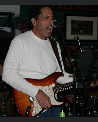 A male musician singing into a microphone while playing a sunburst electric guitar during a live band performance.