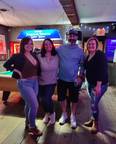 Group of friends smiling in a sports bar next to a pool table with neon beer signs.