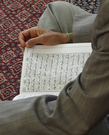 A person sitting on a patterned rug while reading the Holy Quran with Arabic calligraphy.