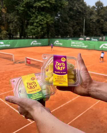 teniso kortų fone rankose laikomi pjaustytų ananasų bei melionų indeliai