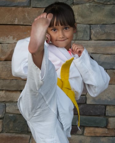 A young girl in a white karate gi and yellow belt performing a high front kick in martial arts class.