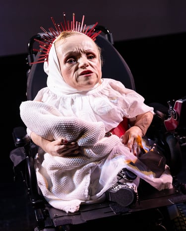 A white woman of very short stature in an electric wheelchair wearing white, with a crown of spikes