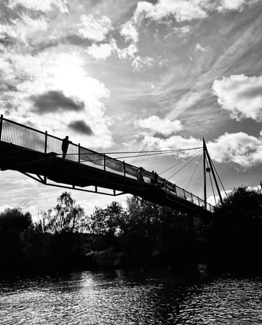 Black and white silhouette of a suspension bridge over a river with a cloudy sky backdrop.