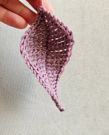 Hand holding a small purple crochet leaf pattern made from wool yarn against a neutral fabric background.