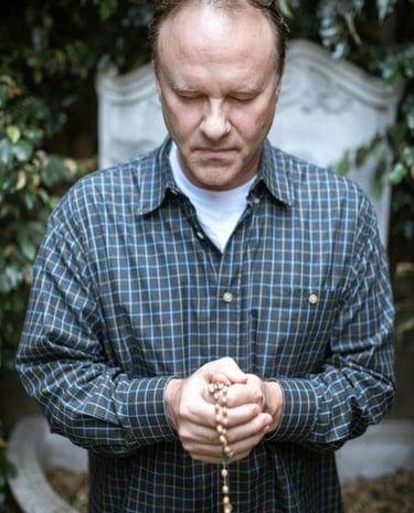 Individual holding a rosary with bowed head in front of a stone monument, surrounded by greenery.