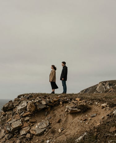 A couple standing quietly together on a windswept coastal cliff, muted earthy tones, overcast sky, cinematic wide-angle storytelling.