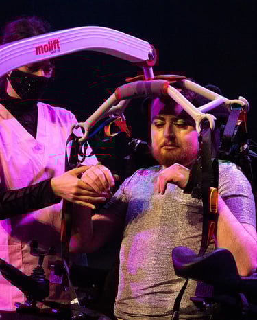 A woman in pink scrubs helps an electric wheelchair user into a hoist and fluffy handcuffs