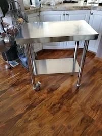 A compact stainless steel table with a bottom shelf and castor wheels sitting on a wooden floor in a home kitchen.