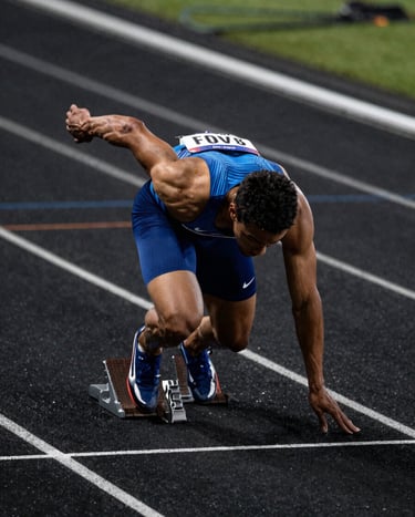 A dramatic, high-shutter speed photograph of a North American sprinter exploding out of starting blocks on a sleek, dark-surfaced track. Cinematic lighting highlights the muscle definition and water droplets, with a color palette of deep black and electric blue.