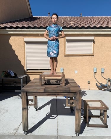 A woman in a blue floral cheongsam dress stands on a tiered wooden table platform outdoors.