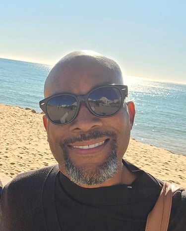 Smiling man wearing sunglasses on a sunny beach with blue ocean water in the background.