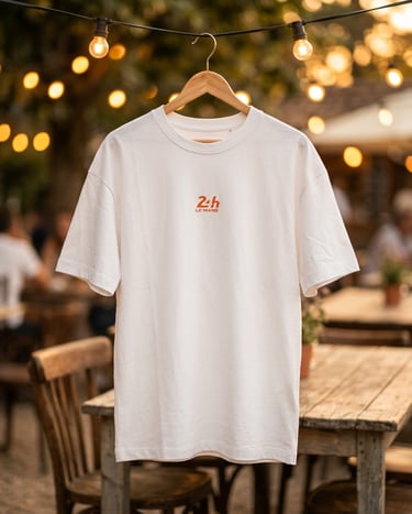 White 24h Le Mans graphic t-shirt hanging outdoors on a wooden hanger with warm string lights.