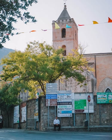 Elderly man in Oliena with street signs and Santa Maria Assunta church tower.