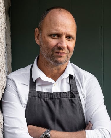Professional chef wearing a white shirt and black apron leaning against a stone wall.