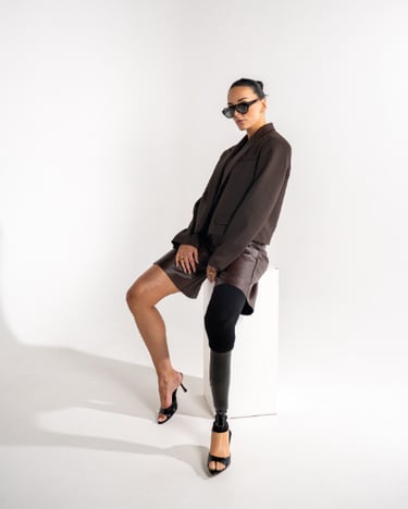 A stylish woman with a prosthetic leg wears a brown blazer and shorts with black sunglasses.