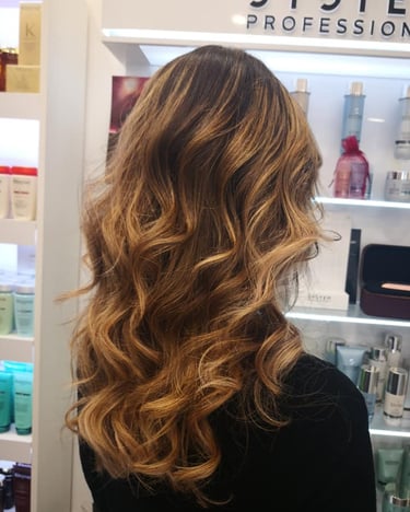 Back view of a woman with long wavy brunette hair and honey blonde balayage highlights in a hair salon.