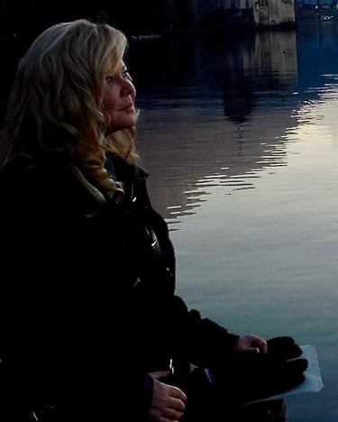 A blonde woman looking at a peaceful sunset reflection on a lake near a historic castle.