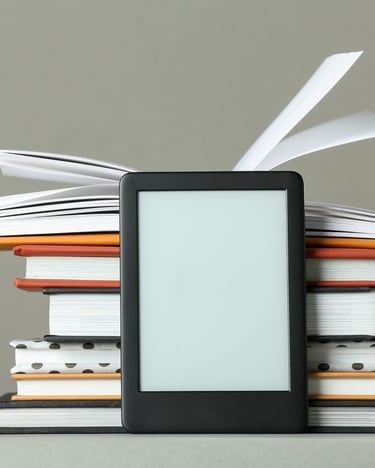 An e-reader with a blank screen leans against a stack of hardcover books with fluttering pages.