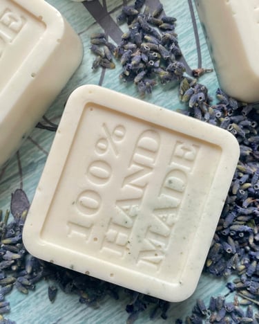 Square white 100% handmade soap bars with embossed text and dried lavender buds on a blue surface.