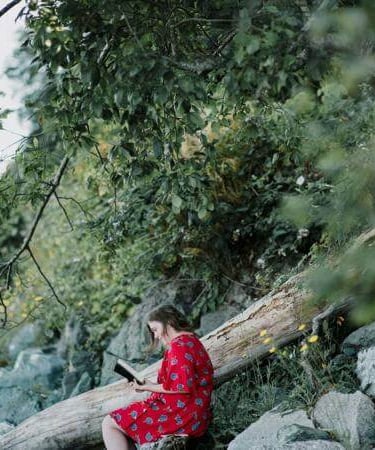 Person in red floral dress reading on a rock in a lush forest setting.