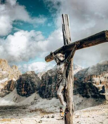 Weathered wooden crucifix with Jesus statue set against snow-capped mountains and cloudy sky.
