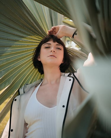 Portrait of Yulia standing outdoors beneath palm leaves, wearing a light jacket and tank top in natural light.