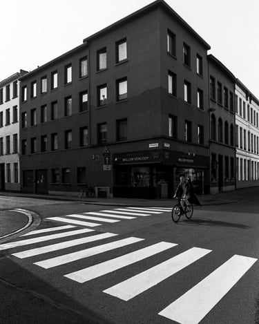 Black and white street photography of a cyclist crossing a zebra walk near a corner building in Antwerp.