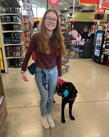 a student dog trainer smiles proudly as her dog holds an object in the grocery store
