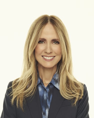 Professional headshot of a smiling blonde woman in a navy blazer and blue patterned shirt.