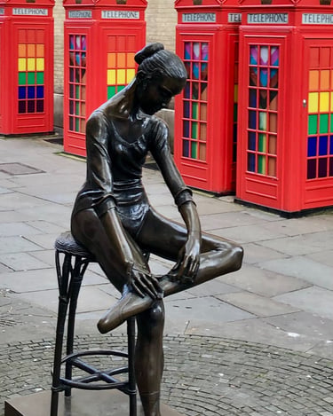 Bronze ballet dancer statue in London with red telephone boxes and colorful glass windows.