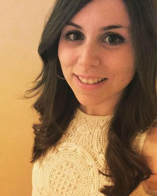 A smiling young woman with long brunette wavy hair wearing an elegant white lace sleeveless dress.