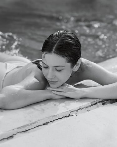Black-and-white portrait of Anna lying by the pool edge with her upper body uncovered, resting her head on her arms.