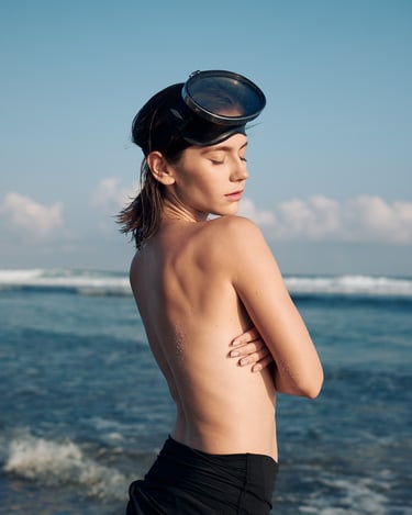 Portrait of Anna standing by the sea with her upper body uncovered, wearing diving goggles and photographed from behind.