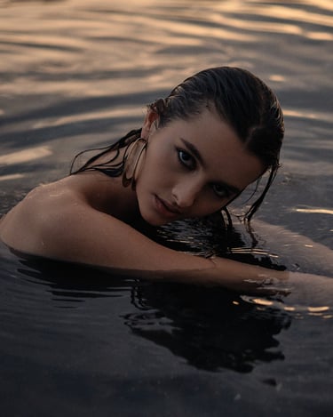 Portrait of Diana partially submerged in water, leaning forward and looking toward the camera in natural light.