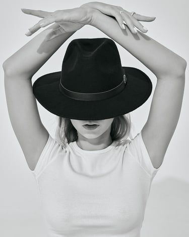 Black-and-white portrait of Kat wearing a hat and white crop top, posing with arms raised above her head.